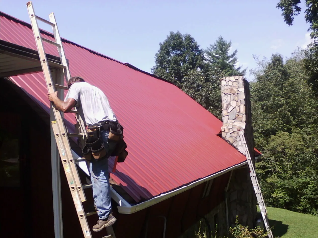 Roofer on a ladder beside a red metal roof and stone chimney — Soffit & Fascia Repair by Roofing Brothers in Bent Mountain